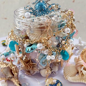 Può includere: Un barattolo trasparente pieno di conchiglie, perle e gioielli dorati. Un ornamento blu a forma di polpo è posto sopra il barattolo. Conchiglie e gioielli aggiuntivi sono sparsi intorno al barattolo, creando un tema costiero.