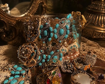 May include: A collection of vintage jewelry, including rings, necklaces, and earrings, in a glass jar. The jewelry features gold-toned metal and turquoise-colored stones. Some pieces have colorful gemstones and pearl accents. The items are displayed on a lace cloth.