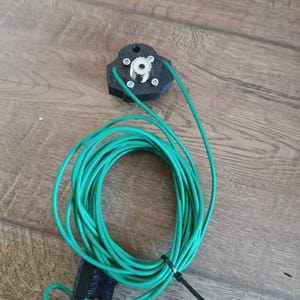 Puede incluir: Un componente electrónico negro, octogonal, con un conector plateado y cables verdes. Los cables están enrollados y asegurados con una brida negra. El componente está sobre una superficie de madera.