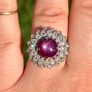 May include: A silver-toned ring featuring a large, round, deep magenta star gemstone at the center. The stone is surrounded by a double halo of small, clear, sparkling stones set in a floral-inspired metal arrangement with intricate detailing.