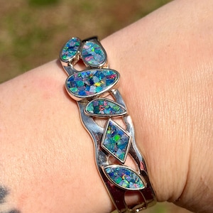 May include: Silver bracelet with inlaid, iridescent opal stones in various shapes. The bracelet has a wavy design with oval, teardrop, and diamond-shaped opal accents. The opals display a mosaic of blues, greens, and hints of other colours.