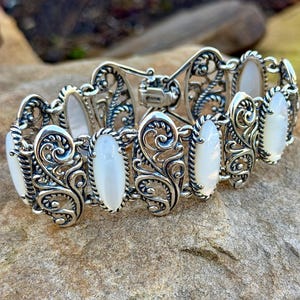 May include: Silver bracelet with oval, white gemstones and intricate, swirling filigree patterns. The bracelet is made up of several hinged segments. It is displayed on a textured, light brown stone surface.