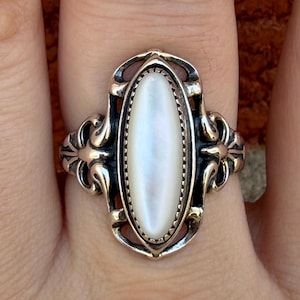 Könnte beinhalten: Ein silberner Ring mit einem ovalen, schillernden Perlmuttstein. Der Ring hat ein verziertes Filigran-Design mit dekorativen Verzierungen an den Seiten. Die Fassung ist detailliert und im Vintage-Stil gehalten.