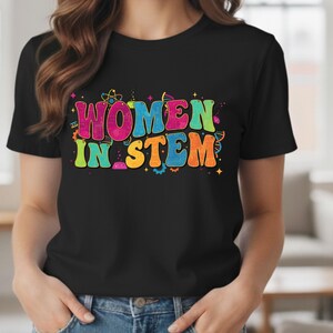 May include: Black t-shirt with the text "WOMEN IN STEM" in colorful, retro-style lettering. The design includes science-themed graphics like atoms and DNA strands. The shirt is short-sleeved and has a crew neck.