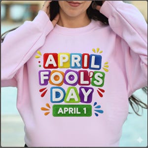 Peut inclure: Un sweat-shirt rose clair avec l'inscription "APRIL FOOL'S DAY APRIL 1" en lettres colorées. Le motif comprend des accents jaunes, rouges, bleus, verts et violets. Le sweat-shirt est un vêtement décontracté.