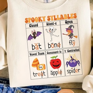 May include: A cream-coloured sweatshirt with a Halloween-themed educational graphic. The design includes the text "SPOOKY SYLLABLES" and illustrations of a bat, skeleton, witch, candy corn, apple, and spider, each paired with a word.