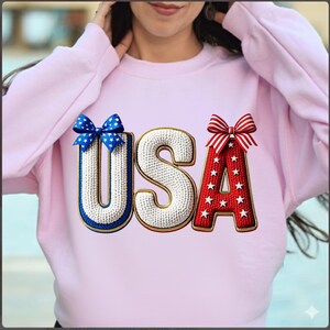 Peut inclure: Un sweat-shirt rose clair avec le mot "USA" dans un design patriotique. Le "U" est bleu avec un nœud, le "S" est blanc et le "A" est rouge avec des étoiles et un nœud. Le sweat-shirt est de style col rond.