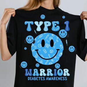 May include: Black t-shirt with a blue smiley face design and the words "TYPE 1 WARRIOR DIABETES AWARENESS" in blue. The smiley face is filled with medical-themed icons. Small smiley faces are scattered around the design.