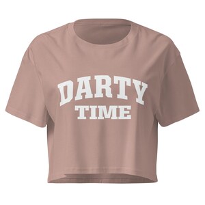 Può includere: Un crop top rosa polveroso con la scritta bianca "DARTY TIME" in un carattere audace e universitario. La maglietta a maniche corte è realizzata in un materiale morbido e ha una vestibilità rilassata.