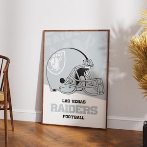 Op de afbeelding: Ingelijste poster met een grijze voetbalhelm met een zwart gezichtsmasker. De poster heeft de woorden "Las Vegas Raiders Football" in grijs en wit. De achtergrond is lichtgrijs en wit. De poster zit in een bruine lijst.