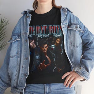 May include: Black t-shirt featuring the text "THE BAT BOYS" above three male character portraits. The names "Feyre", "Rhysand", and "Cassian" are below the text. The shirt is worn with a blue denim jacket.