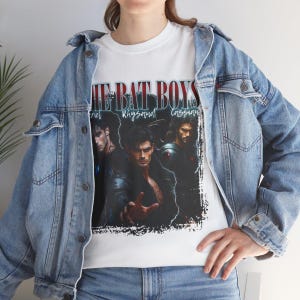 May include: White t-shirt featuring a graphic of three men with the text "THE BAT BOYS" above them. The names "Feyre", "Rhysand", and "Cassian" are printed below the men. The shirt is worn with a light blue denim jacket.
