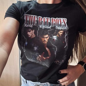 May include: Black t-shirt featuring the text "THE BAT BOYS" above three male character portraits. The names Azriel, Rhysand, and Cassian are printed below the text. The shirt is worn with blue jeans.