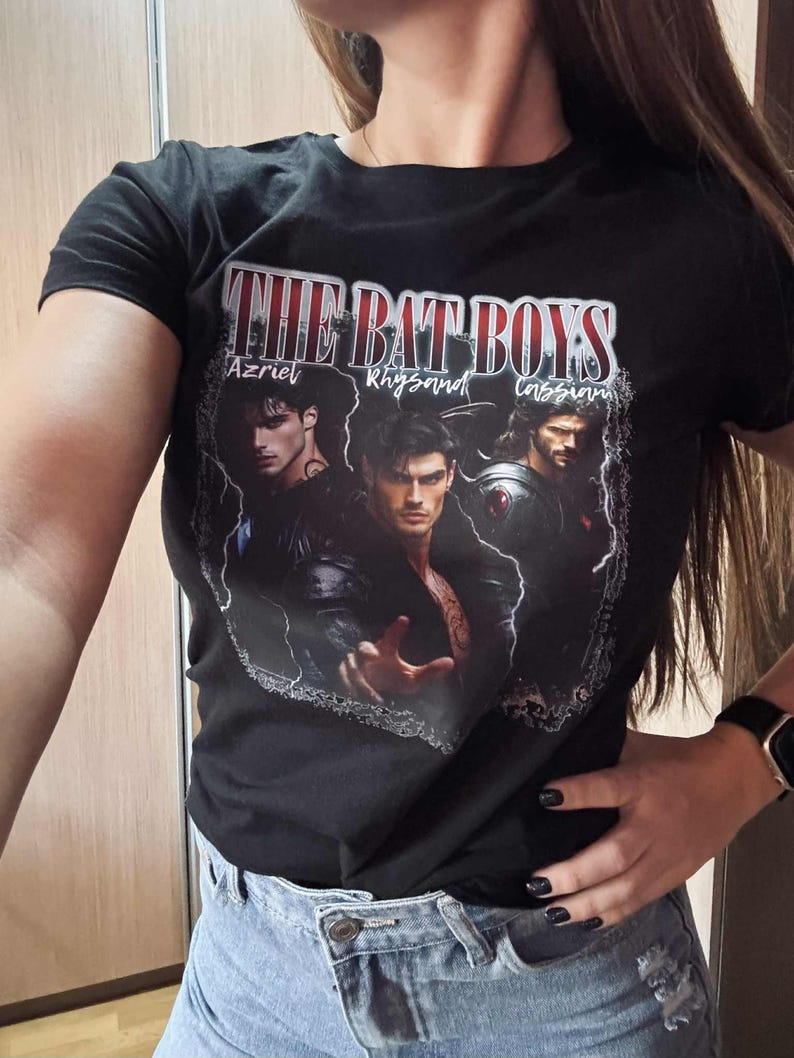 May include: Black t-shirt featuring the text "THE BAT BOYS" above three male character portraits. The names Azriel, Rhysand, and Cassian are printed below the text. The shirt is worn with blue jeans.