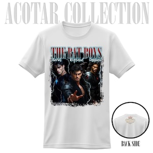 May include: White t-shirt featuring the text "THE BAT BOYS" above a graphic of three male characters. The names Azriel, Rhysand, and Cassian are below the text. The back of the shirt is visible in a small circle.