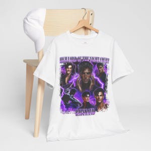 May include: White t-shirt featuring a graphic design with multiple images of a man, purple lightning bolts, and the text "HIGH LORD OF THE NIGHT COURT" and "RHYSAND". The shirt is hanging on a wooden hanger.