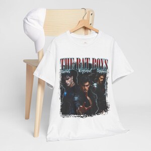 May include: White t-shirt featuring the text "THE BAT BOYS" above a graphic of three men. The names Azriel, Rhysand, and Cassian are below the text. A white baseball cap rests on a wooden chair.