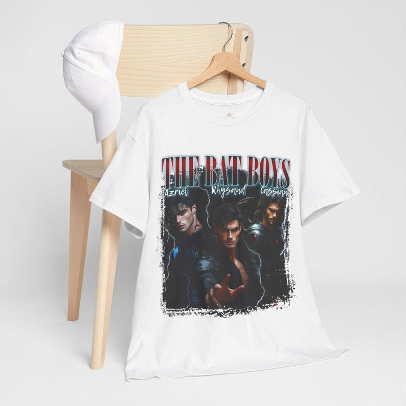May include: White t-shirt featuring the text "THE BAT BOYS" above a graphic of three men. The names Azriel, Rhysand, and Cassian are below the text. A white baseball cap rests on a wooden chair.