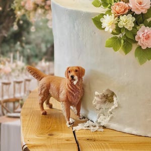 May include: A light-coloured cake decorated with pastel flowers and a small, brown dog figurine. The cake has a hole in the side with frosting around it. The cake sits on a wooden surface.