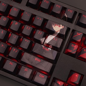 May include: A black mechanical keyboard with red backlit keys. The Enter key features a custom keycap with an anime-style illustration. The keyboard has standard QWERTY layout with function keys and control keys.