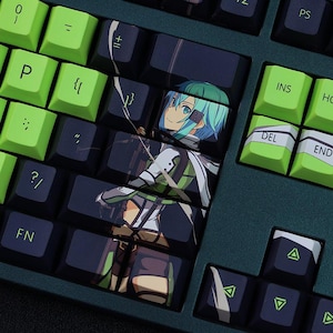 May include: Close-up of a mechanical keyboard with green and black keycaps. Some keycaps are green with white lettering, while others are black with a character illustration. The keyboard has a dark green frame.