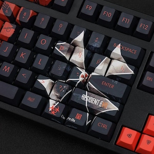 Könnte beinhalten: Eine schwarze mechanische Tastatur mit individuellen Tastenkappen. Die Tastenkappen zeigen ein Resident Evil-Thema mit dem Umbrella Corporation-Logo und anderen spielbezogenen Grafiken. Die Tasten haben rote und weiße Buchstaben.