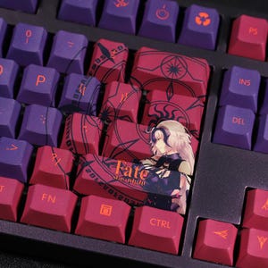 May include: A close-up of a mechanical keyboard with custom keycaps. The keycaps are a mix of purple and red, with various symbols and the text "Fate stay night" printed on them. The keyboard is black.