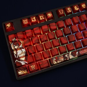 May include: A black mechanical keyboard with red keycaps. Some keycaps feature anime character illustrations and symbols. The keyboard has a Fairy Tail theme, with the logo visible on the spacebar. The keys are backlit with a rainbow glow.