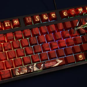 May include: A mechanical keyboard with red keycaps and a black frame. The keycaps have a marbled effect and feature custom designs and symbols. The space bar displays the text "FAIRY TAIL" with anime characters. The keyboard is lit with a colorful backlight.