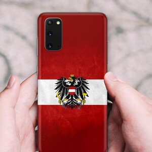 May include: A smartphone case featuring the Austrian flag design with a horizontal red and white tricolor pattern and a central black eagle emblem holding a golden hammer and sickle, topped with a mural crown, set against a textured red background.