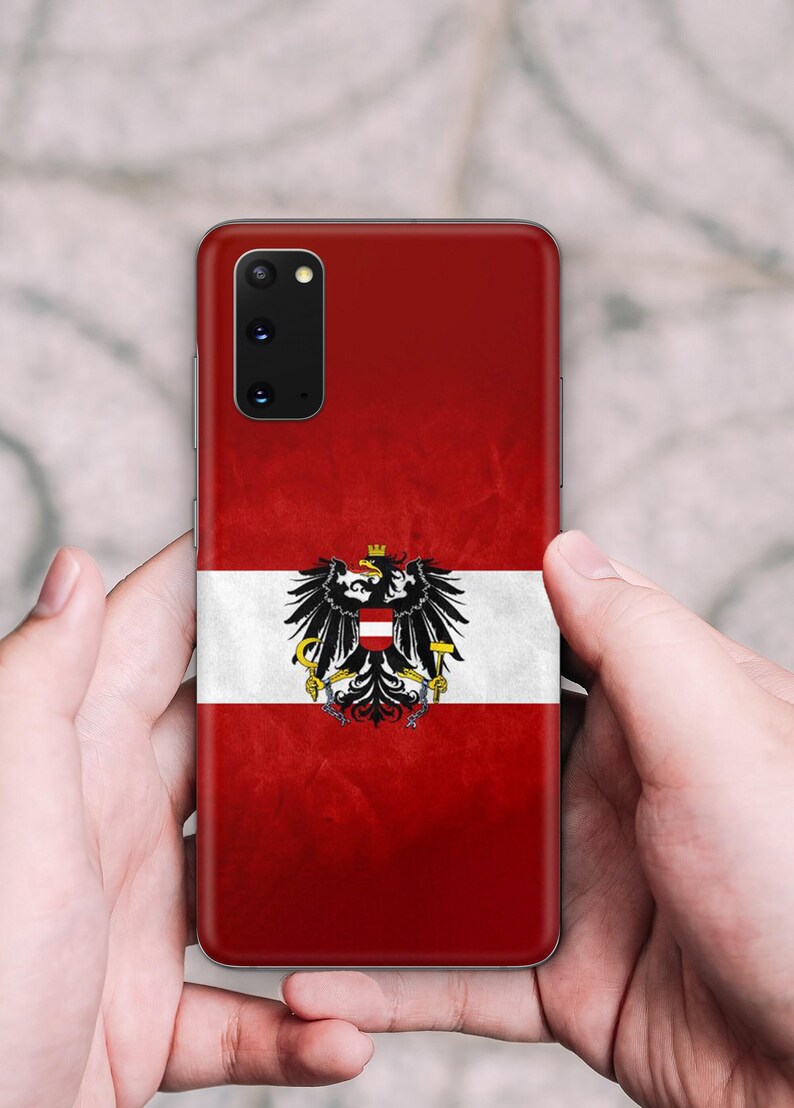 May include: A smartphone case featuring the Austrian flag design with a horizontal red and white tricolor pattern and a central black eagle emblem holding a golden hammer and sickle, topped with a mural crown, set against a textured red background.