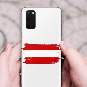 May include: A white smartphone case featuring a horizontal brushstroke design in red and white, mimicking the flag of Austria. The case has a rectangular camera cutout in the upper left corner with multiple lenses and a small circular flash component.