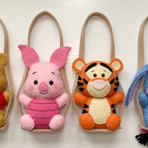 Op de afbeelding: Vier vilten karaktertassen met Winnie de Poeh, Knorretje, Tijger en Iejoor. Elk pluche karakter zit in een beige vilten schommel met een bijpassende handgreep. De karakters zijn in verschillende kleuren, waaronder geel, roze, oranje en blauw.
