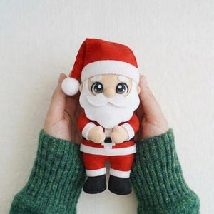 May include: A plush Santa Claus doll with a red hat and suit, white beard, and black boots. The doll is held by two hands wearing a green jumper. The doll is a festive Christmas decoration.