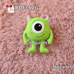 May include: A lime green felt monster with one large black and white eye, small white horns, and a smiling mouth. The monster has arms and legs and the text "MagixFelt" is visible. The background is a pink, fluffy surface.