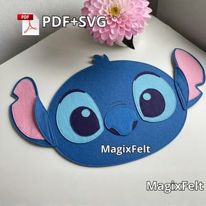 May include: A blue felt placemat shaped like a cartoon character's face, with large eyes, ears, and a nose. The character's face is blue, with light blue and purple accents. The ears are pink. The text "MagixFelt" is visible.