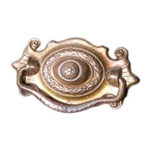 Puede incluir: Tirador de cajón de latón antiguo con un centro ovalado con un diseño floral y un borde decorativo. El tirador tiene un asa curvada y detalles ornamentados en los laterales.