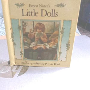 Könnte beinhalten: Ein antikes Buch mit dem Titel "Little Dolls" von Ernest Nister. Das Cover zeigt eine Illustration eines Kindes und den Text "An Antique Moving Picture Book". Das Buch hat einen cremefarbenen Einband mit dekorativen floralen Akzenten.