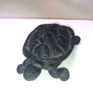 May include: A dark gray turtle figurine with a textured shell and detailed legs. The figurine is made of a solid material and sits on a white surface. The turtle's shell has a patterned design.