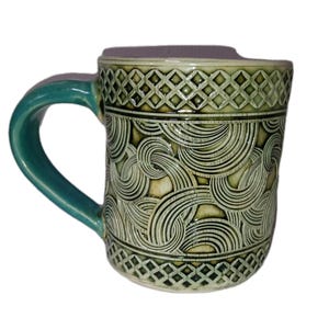 May include: A ceramic mug with a teal handle and a detailed, carved design. The mug features a repeating pattern of swirling lines and diamond shapes in shades of green and cream. The mug is approximately 4 inches tall.