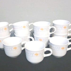 Puede incluir: Un juego de tazas de café de cerámica blanca con asas. Cada taza presenta un diseño floral en naranja y gris. Las tazas están apiladas y dispuestas sobre una superficie oscura, ideal para la cocina o el comedor.