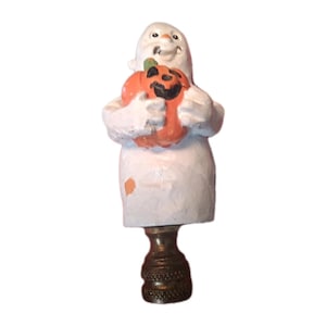 May include: A white ghost figurine holding an orange jack-o'-lantern with a black carved face. The ghost has a smiling expression and is mounted on a dark metallic lamp finial base with a textured, knurled grip pattern at the bottom.