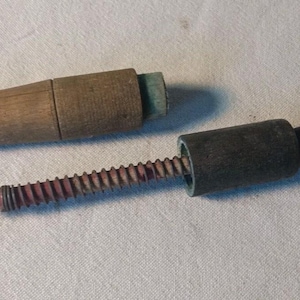 Könnte beinhalten: Ein altes Werkzeug aus Holz, das aus zwei separaten Teilen besteht. Ein Teil ist ein zylindrischer Holzgriff mit einer langen, gewindeten Metallschraube. Der zweite Teil ist ein hohles Holzgehäuse, das über die Gewindeschraube passt.
