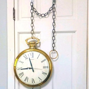 May include: A large, gold-colored pocket watch clock hanging from a black chain on a white door. The clock face has black Roman numerals and hands. The clock is suspended by a metal bracket.