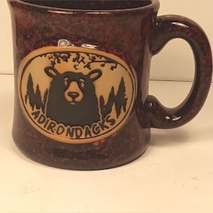 May include: A dark brown speckled ceramic mug featuring a central oval emblem. The emblem displays a black bear silhouette against a forest background with the text ADIRONDACKS printed in black lettering along the bottom curve of the oval.