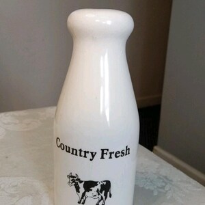 Op de afbeelding: Een witte keramische melkfles met een smalle hals en een afgeronde rand. Op de voorzijde van de fles staat de zwarte tekst "Country Fresh" gedrukt boven een zwart-witte grafische illustratie van een koe die in profiel staat.