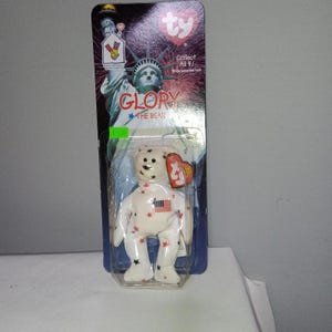 Pode incluir: Um urso de peluche Ty Beanie Baby branco chamado Glory na sua embalagem original. O urso apresenta padrões de estrelas vermelhas e azuis e um pequeno emblema da bandeira americana no peito. O cartão exibe a Estátua da Liberdade.