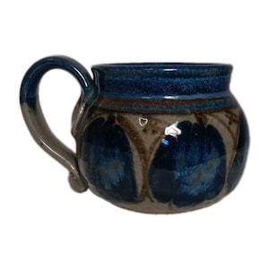 May include: A rounded ceramic mug featuring a glossy deep blue glaze on the upper rim and handle, transitioning into a beige body decorated with repeating dark blue circular patterns separated by brown horizontal bands and textured accents.