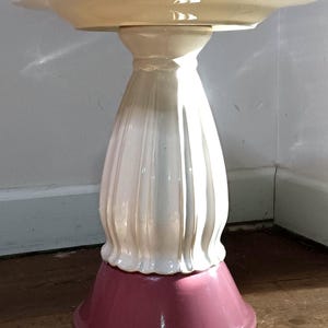 Puede incluir: Un pedestal decorativo de cerámica con una parte superior festoneada y una base acampanada. La parte superior es de color crema claro, el pedestal es blanco y la base es de color rosa intenso. El pedestal mide unos 30 cm de altura.