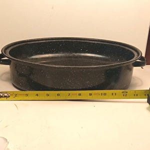 May include: An oval black roasting pan with white speckles and two side handles. A yellow measuring tape is positioned in front of the pan, showing a length of approximately 14 inches from the left edge to the right handle.